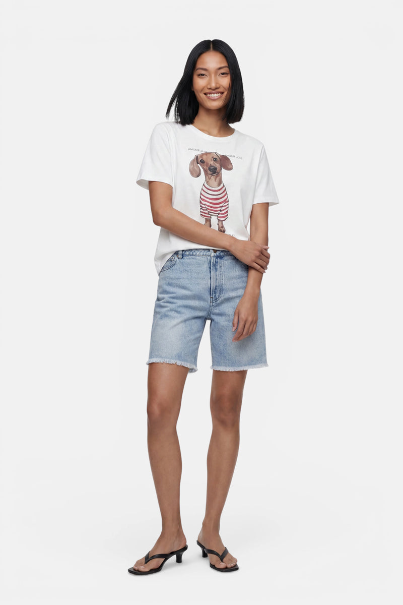 Vor einem schlichten weißen Hintergrund steht eine lächelnde Frau. Sie trägt ein T-Shirt mit einem „Princess Goes Hollywood“-Motiv, das einen Dackel zeigt, Jeansshorts mit leichter Waschung und ausgefranstem Saum sowie schwarze Sandalen mit Absatz. Ihre Arme sind verschränkt, und ihr schulterlanges Haar ist glatt.