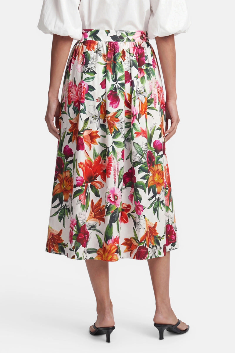 Eine Person steht mit dem Rücken zur Kamera und trägt eine weiße Bluse sowie einen hoch taillierten Midirock mit Blumenmuster aus Baumwolle von Princess Goes Hollywood in leuchtendem Rot, Orange, Pink und Grün auf weißem Hintergrund. Sie trägt schwarze Sandalen mit Absatz vor einem schlichten weißen Hintergrund.