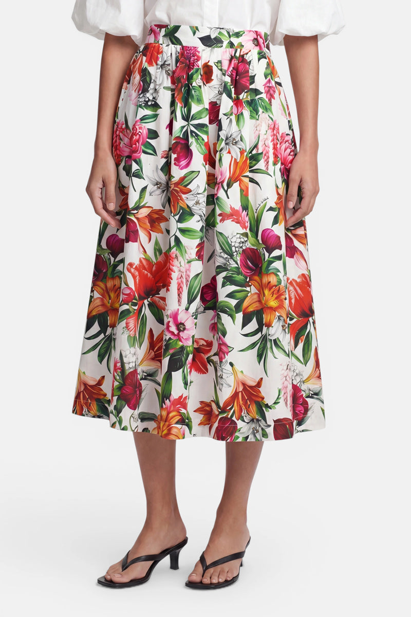Eine Frau trägt einen weiten, plissierten Midirock aus Baumwolle mit buntem Blumenmuster von Princess Goes Hollywood. Dazu kombiniert sie eine weiße Bluse mit Puffärmeln und schwarze Sandaletten vor einem schlichten, weißen Hintergrund.