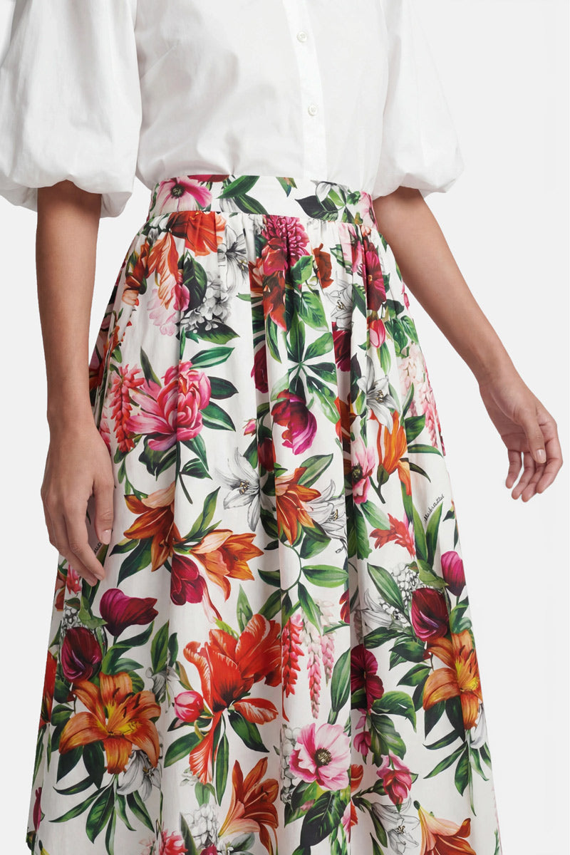 Eine Frau trägt eine weiße Bluse mit Knöpfen und Puffärmeln sowie einen hoch taillierten Midirock der Marke „Princess Goes Hollywood“ aus Baumwolle, der mit einem leuchtenden Blumenprint in Rot, Orange, Rosa und Grün auf weißem Hintergrund verziert ist. Zu sehen sind nur ihr Oberkörper und ihre Oberschenkel.