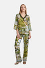 Eine Frau mit langen blonden Haaren steht mit dem Blick nach vorne und trägt ein grün-weißes Outfit mit Safari-Print von Princess Goes Hollywood. Das Outfit besteht aus einem locker sitzenden Oberteil mit V-Ausschnitt und ellbogenlangen Ärmeln sowie einer weit geschnittenen Seidenhose mit elastischem Bund, dazu trägt sie Sandalen mit Absatz.