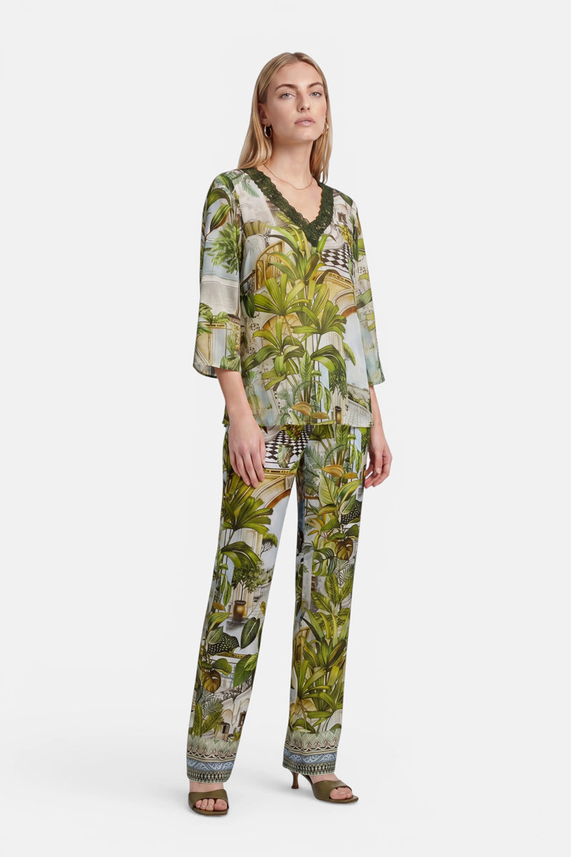 Eine Frau präsentiert ein locker sitzendes, aufeinander abgestimmtes zweiteiliges Outfit in tropischem Grün mit Safari-Print von Princess Goes Hollywood. Die Seidenbluse hat Dreiviertelärmel und einen V-Ausschnitt, der mit dunklem Stoff eingefasst ist. Vor einem weißen Hintergrund kombiniert sie dazu Pumps mit offener Spitze.