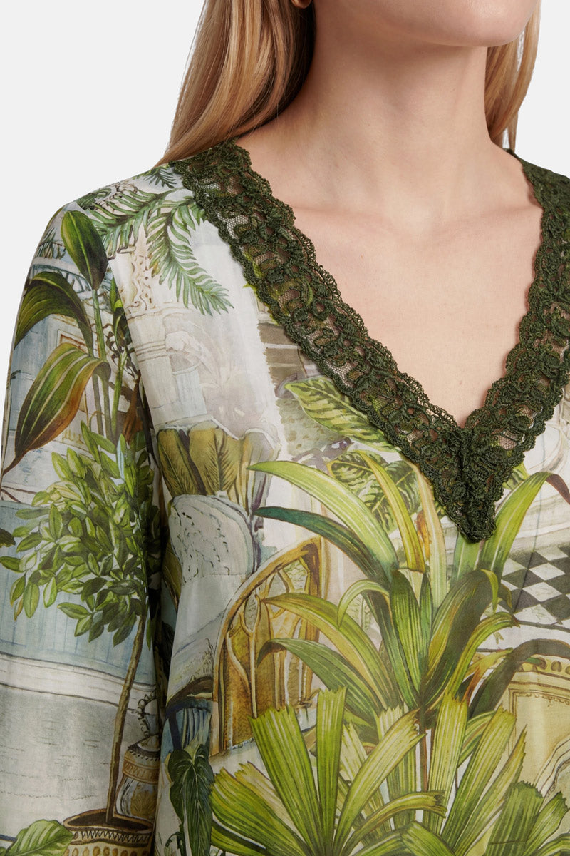 Eine Frau trägt eine Seidenbluse von „Princess Goes Hollywood“ mit V-Ausschnitt und dunkelgrüner Spitzenborte, verziert mit einem farbenfrohen Botanik- und Safari-Print, der üppige Pflanzen und architektonische Motive zeigt. Ihr langes blondes Haar umrahmt ihr Gesicht, das teilweise außerhalb des Bildausschnitts liegt.