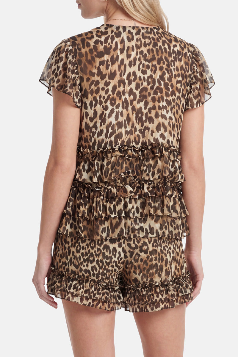 Eine Frau steht mit dem Rücken zur Kamera und trägt einen Overall der Marke „Princess Goes Hollywood“ mit Leopardenmuster, mehrlagigen Rüschen an der Taille und Shorts. Der transparente, leichte Stoff und der V-Ausschnitt verleihen dem Outfit Eleganz. Ihre Arme liegen an den Seiten, sodass nur ihr oberer Rücken und ihre Beine zu sehen sind.