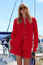 Eine Frau mit blonden Haaren und einer großen Sonnenbrille steht in einem Yachthafen neben Segelbooten. Sie trägt eine übergroße, leuchtend rote Baumwollmusselin-Hemdbluse aus der „Princess Goes Hollywood Limited Edition“ sowie dazu passende Shorts und hält unter dem strahlend blauen Himmel eine gestreifte Tasche in der Hand.