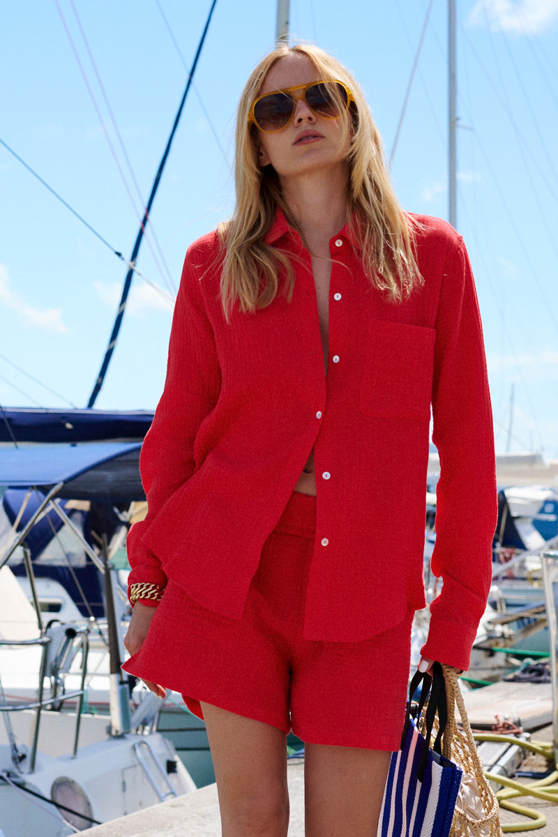 Eine Frau steht auf einem Steg vor einigen Booten und trägt ein lockeres, leuchtend rotes Baumwollmusselin-Hemd mit Knöpfen aus der „Princess Goes Hollywood“-Limited-Edition sowie dazu passende Shorts. Sie hat langes blondes Haar, trägt eine große braune Sonnenbrille und hält eine blau gestreifte Tasche sowie eine goldene Tragetasche in der Hand.