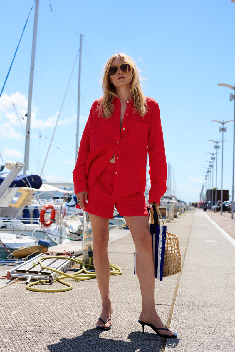 Eine selbstbewusste Frau mit langen blonden Haaren und Sonnenbrille steht auf einem Steg im Yachthafen und trägt eine knallrote „Limited Edition“-Hemdbluse sowie dazu passende Shorts von „Princess Goes Hollywood“, dazu eine gestreifte Strohtasche und schwarze Sandalen mit Absatz.