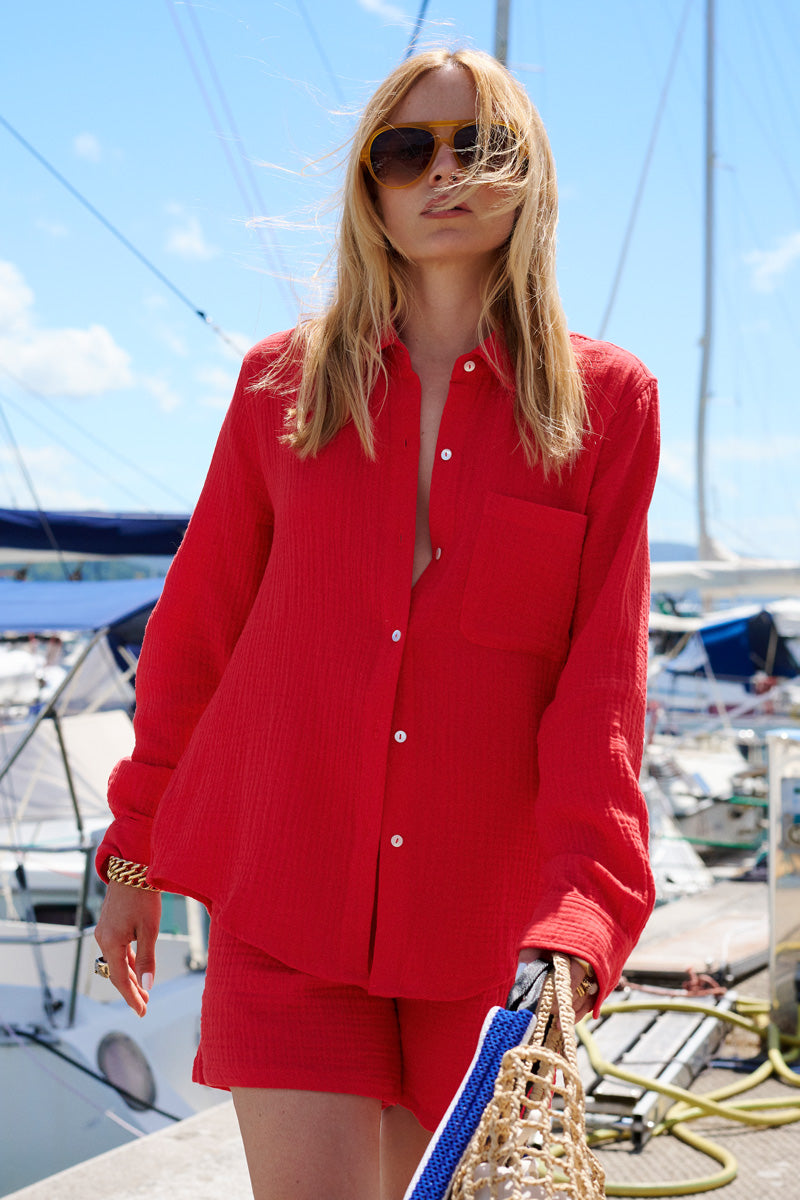 Eine Frau in einer leuchtend roten Hemdbluse in limitierter Auflage und passenden Shorts von Princess Goes Hollywood steht auf einem Steg im Yachthafen, eine geflochtene Tasche in der Hand und eine große Sonnenbrille auf der Nase. Segelboote, aufgerollte Seile und ein teilweise bewölkter Himmel umgeben sie.