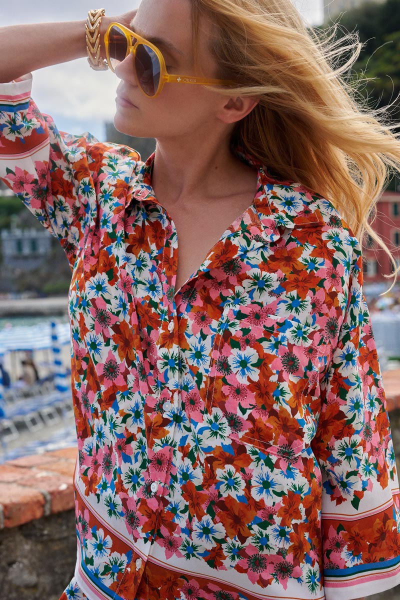 Eine Person mit langen blonden Haaren trägt eine übergroße gelbe Sonnenbrille und eine farbenfrohe Princess Goes Hollywood Limited Edition Hemdbluse mit roten, rosa und blauen Blumen. Sie steht im Freien an einem Ziegelgeländer mit Wasser und bunten Gebäuden dahinter.