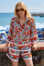 Eine Frau mit langen blonden Haaren trägt eine große orangefarbene Sonnenbrille und ein passendes Princess Goes Hollywood Limited Edition Blumenprint-Shirt und Seidenshorts in Rot, Rosa, Blau und Weiß. Sie steht am Meer, mit gestreiften Regenschirmen und einem Boot hinter ihr.
