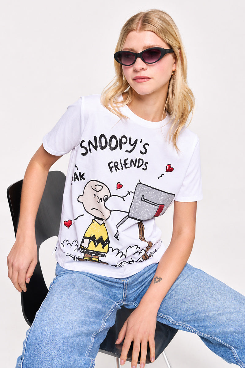 Eine blonde Frau trägt eine Princess Goes Hollywood-Sonnenbrille in limitierter Auflage, ein Snoopy-T-Shirt aus Baumwolle und hellblaue Jeans, während sie auf einem schwarzen Stuhl sitzt. Sie hat ein kleines Herz-Tattoo auf ihrem linken Handgelenk und sieht entspannt aus.