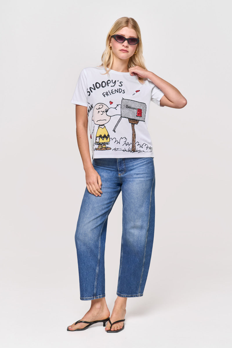 Eine Frau steht vor einem schlichten weißen Hintergrund und berührt ihre Sonnenbrille, während sie blaue Jeans, schwarze Flip-Flops und ein Princess Goes Hollywood Limited Edition Snoopy T-Shirt aus Baumwolle mit Charlie Brown- und Snoopy-Grafiken trägt.
