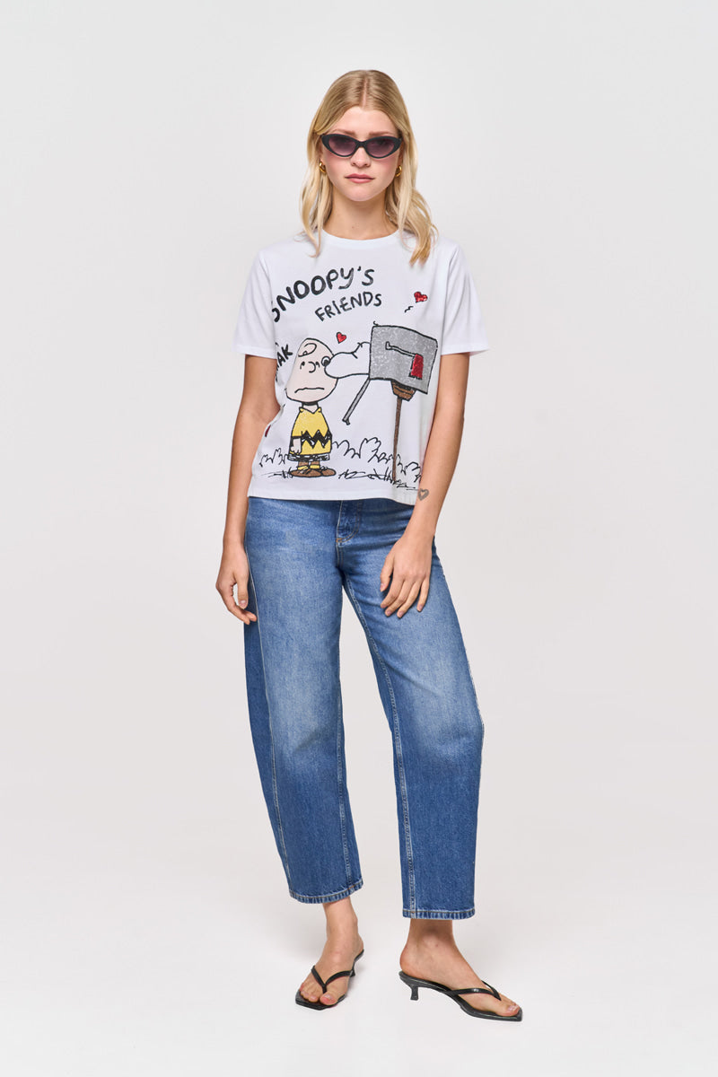 Eine blonde Frau mit rosa Sonnenbrille, blauer Jeans, schwarzen Flip-Flops und einem Princess Goes Hollywood Limited Edition "Snoopy T-Shirt", das Charlie Brown und Snoopy an einem Briefkasten zeigt, steht vor einem schlichten weißen Hintergrund.