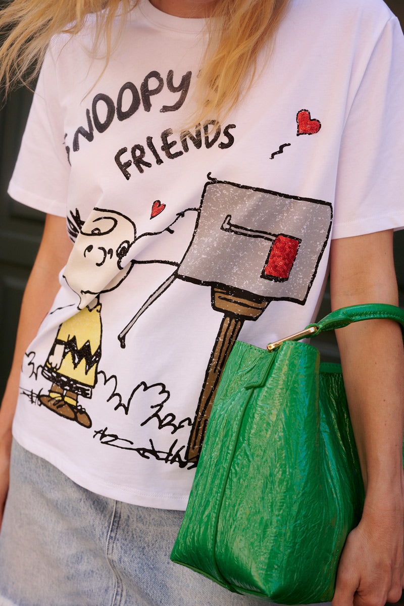 Eine Person trägt ein weißes Snoopy-T-Shirt aus der „Princess Goes Hollywood“-Limited-Edition, auf dem Charlie Brown an einem Briefkasten und rote Herzen abgebildet sind, dazu einen hellblauen Jeansrock und eine leuchtend grüne, strukturierte Handtasche. Zu sehen sind nur der untere Teil ihres Gesichts, ihr Oberkörper und einige blonde Haarsträhnen.