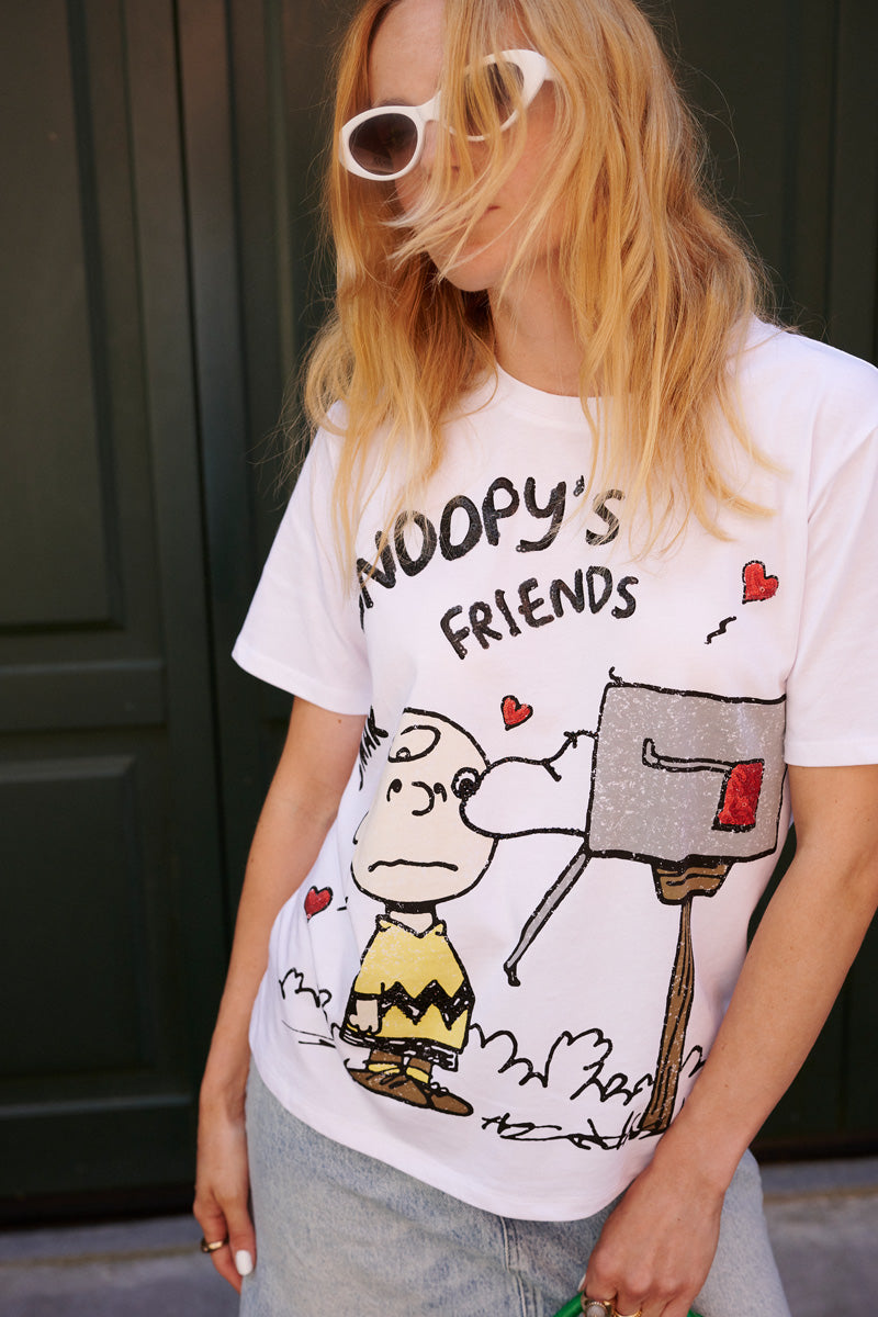 Eine Person mit langen blonden Haaren, einer weißen Sonnenbrille und einem „Princess Goes Hollywood“-Snoopy-T-Shirt in limitierter Auflage, auf dem Charlie Brown abgebildet ist, steht vor einem dunkelgrünen Türflügel neben einem Briefkasten mit roten Herzen und hält etwas Grünes in der Hand.