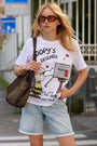 Eine Frau mit langen blonden Haaren trägt eine getönte Sonnenbrille, ein „Princess Goes Hollywood Limited Edition“-Snoopy-T-Shirt aus Baumwolle, helle Jeansshorts und hält eine große geflochtene Tasche in der Hand. Sie steht in der Nähe eines Zebrastreifens und stützt eine Hand in die Hüfte.