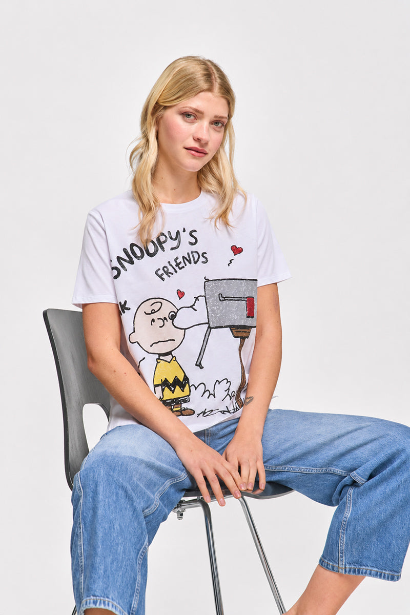 Eine junge Frau mit langen blonden Haaren sitzt auf einem schwarzen Stuhl und trägt ein Princess Goes Hollywood Limited Edition Snoopy T-Shirt aus Baumwolle mit Charlie Brown am Briefkasten, lässige Bluejeans, auf einem schlichten weißen Hintergrund und einen neutralen Gesichtsausdruck.