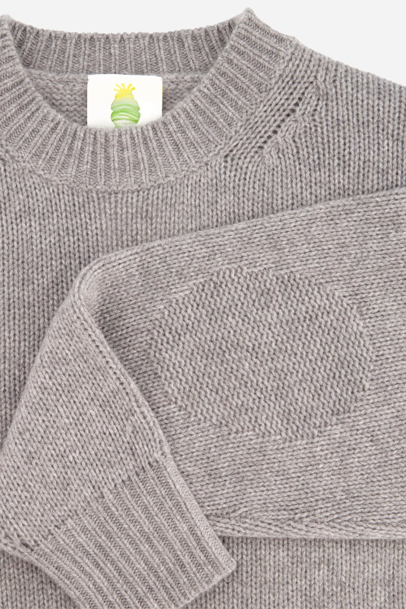 Eine Nahaufnahme eines grauen Kaschmir-Strickpullovers mit strukturierten, ovalen Ellbogenpatches. Der weiche Pullover verfügt über gerippte Bündchen und einen Rundhalsausschnitt. Am Hals ist ein weißes Etikett mit einem abstrakten Logo in Grün und Gelb zu sehen. Der Hintergrund ist einfarbig weiß.