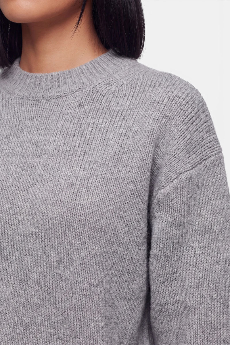 Eine Person, die einen weichen, hellgrauen Kaschmir-Strickpullover mit Rundhalsausschnitt trägt. Das Bild zeigt ihre Schulter, ihren Hals und einen Teil der Kinnlinie. Der Hintergrund ist schlicht weiß, und der Gesamteindruck ist schlicht und gemütlich.