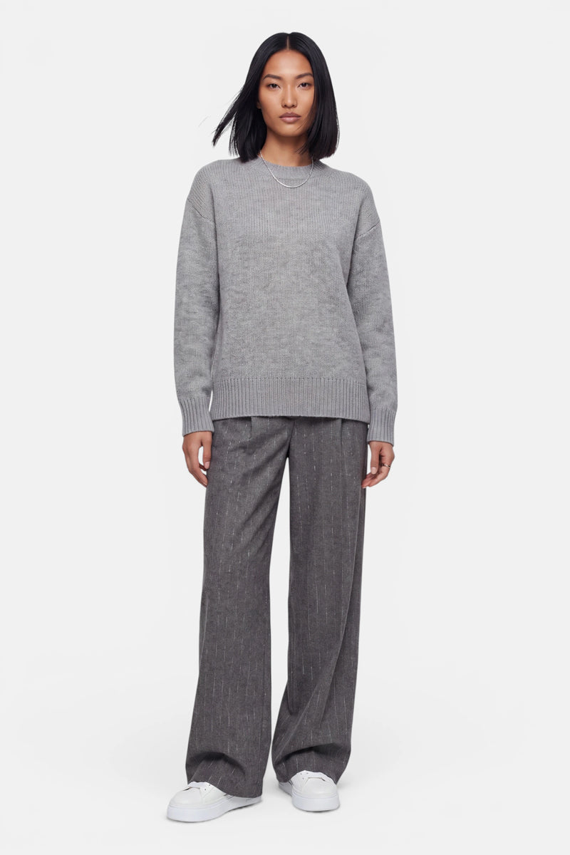 Eine Frau steht mit dem Blick nach vorne und trägt einen lockeren, weichen grauen Strickpullover mit Rundhalsausschnitt, eine graue Nadelstreifenhose mit weitem Bein und weiße Sneaker. Ihr Haar ist glatt und schulterlang. Der Hintergrund ist schlicht weiß. Ihr Gesichtsausdruck ist neutral, und ihre Arme liegen an den Seiten.