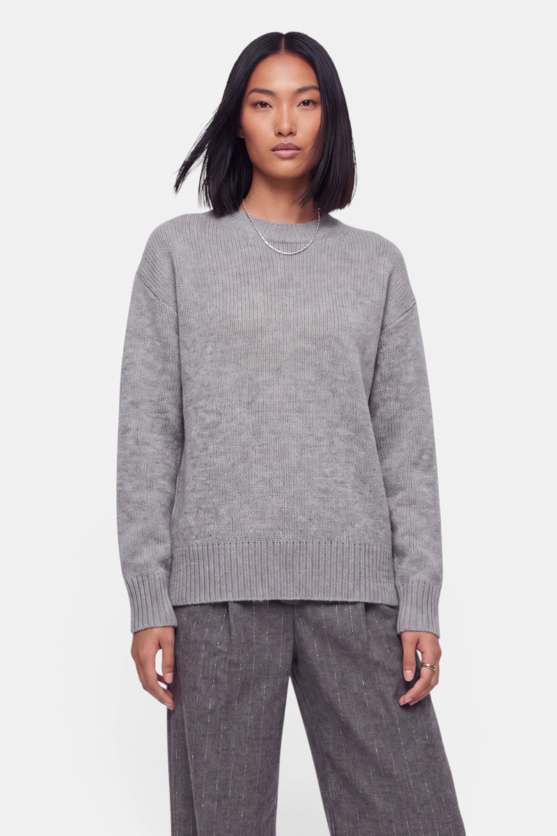 Eine Frau mit glattem schwarzem Haar trägt einen hellgrauen Kaschmir-Strickpullover mit Rundhalsausschnitt und eine locker sitzende graue Nadelstreifenhose. Als Accessoires trägt sie eine schlichte Halskette und einen Ring. Sie steht vor einem schlichten weißen Hintergrund und blickt mit neutralem Gesichtsausdruck direkt in die Kamera.