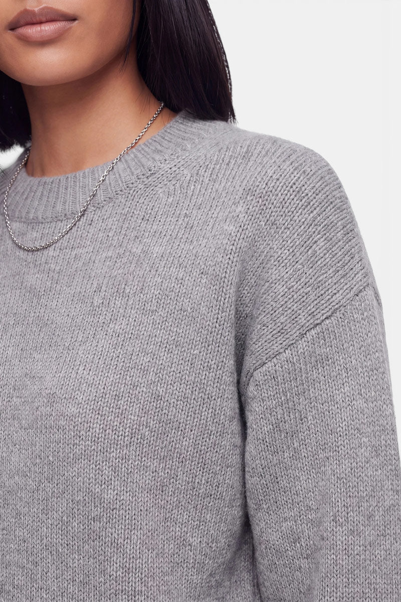 Eine Frau, die einen hellgrauen Kaschmir-Strickpullover mit Rundhalsausschnitt und eine dünne Kettenhalskette trägt, ist von den Schultern aufwärts zu sehen. Ihr glattes, dunkles Haar fällt ihr über die Schultern, und ihre Lippen sind leicht geöffnet. Der Hintergrund ist schlicht weiß.