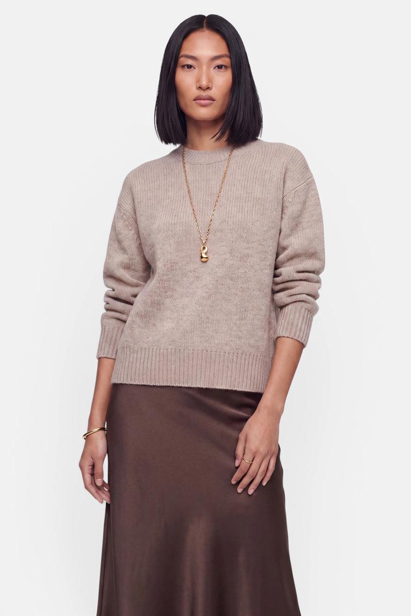 Eine Frau mit glattem schwarzem Haar trägt einen beigen Kaschmir-Strickpullover mit langen Ärmeln und klassischem Rundhalsausschnitt, dazu eine lange goldene Halskette, einen braunen Seiden-Midirock und ein goldenes Armband. Sie steht vor einem schlichten weißen Hintergrund und nimmt eine entspannte, selbstbewusste Haltung ein.