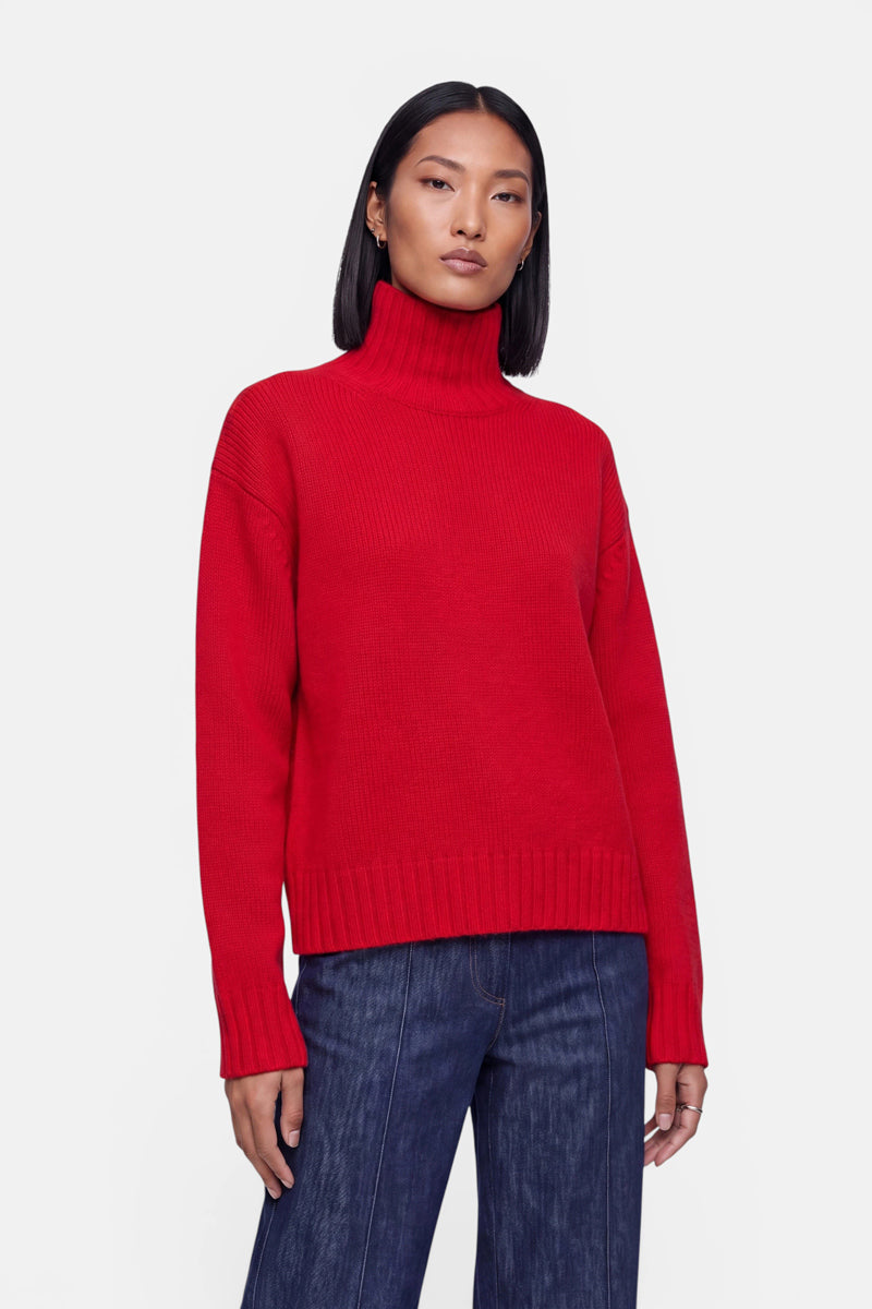 Eine Person mit glattem, schulterlangem schwarzem Haar trägt einen leuchtend roten Kaschmir-Strickpullover mit Stehkragen und dunkelblaue Jeans und steht vor einem schlichten weißen Hintergrund. Der Damenpullover hat eine lockere Passform mit gerippten Bündchen und Saum. Die Person hat einen neutralen Gesichtsausdruck.