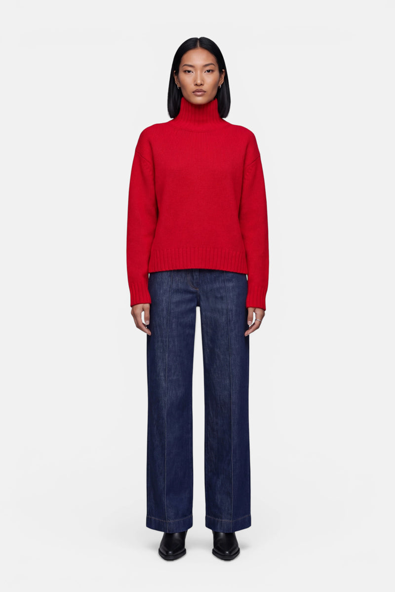 Eine Person steht mit dem Blick nach vorne und trägt einen roten Kaschmir-Strickpullover mit Stehkragen, eine dunkelblaue Jeans mit weitem Bein und schwarze Schuhe. Sie hat glattes, schulterlanges schwarzes Haar und einen neutralen Gesichtsausdruck, vor einem schlichten weißen Hintergrund.