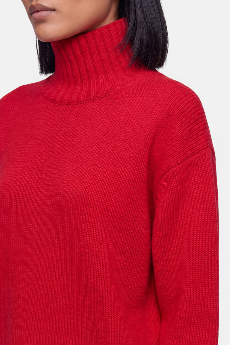 Eine Person mit glattem schwarzem Haar trägt einen leuchtend roten Damenpullover mit geripptem Stehkragen. Das Bild zeigt nur den unteren Teil des Gesichts, den Hals, die Schultern und den Oberkörper vor einem schlichten weißen Hintergrund. Die Struktur des Pullovers und die Strickdetails des Kaschmir-Pullovers sind deutlich zu erkennen.