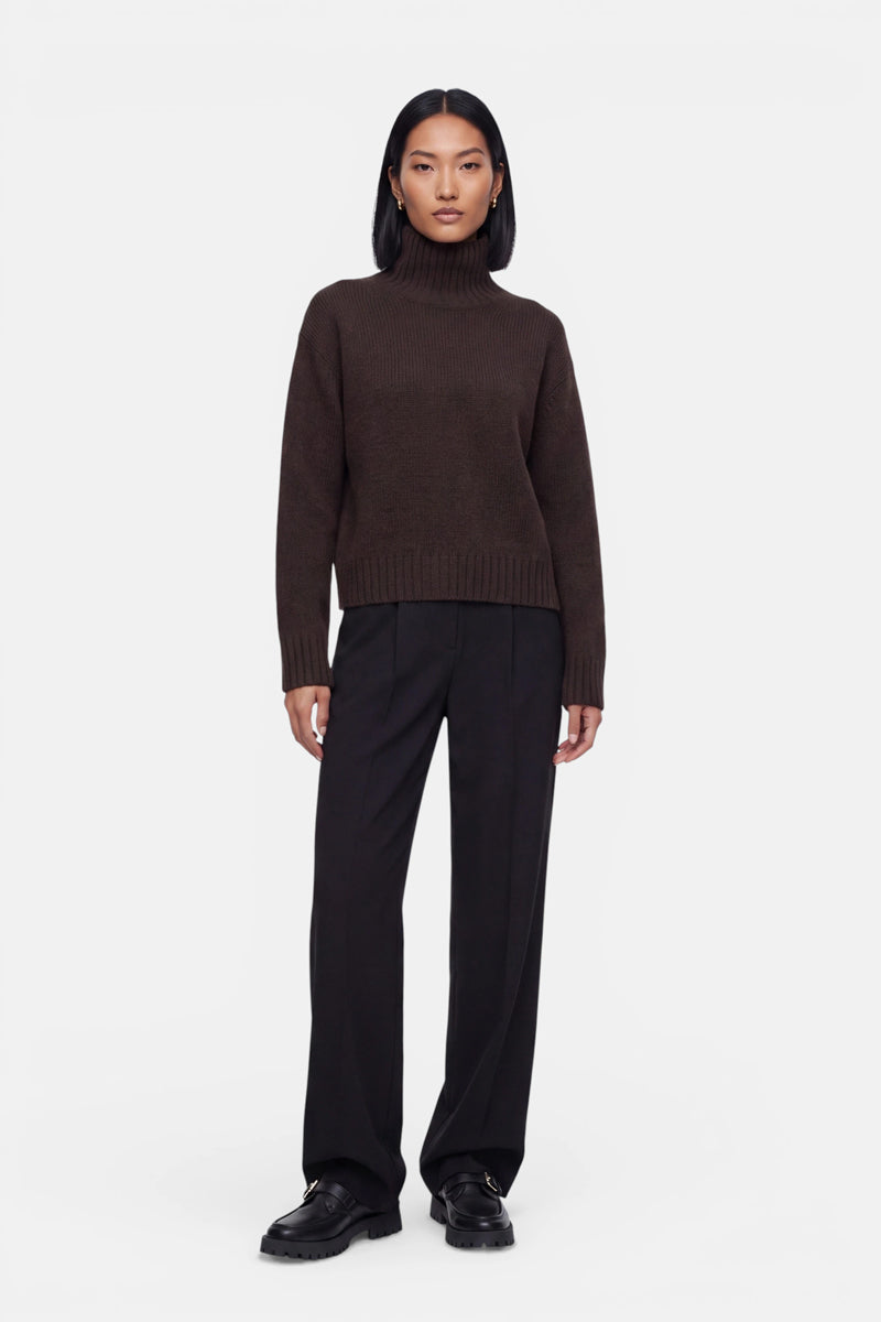 Eine Person steht vor einem weißen Hintergrund und trägt einen braunen Kaschmir-Strickpullover mit Stehkragen, eine schwarze Hose mit weitem Bein und schwarze Schuhe. Die Arme liegen an den Seiten, das glatte, schulterlange Haar fällt locker herab, und die Person blickt mit ausdruckslosem Gesicht direkt in die Kamera.