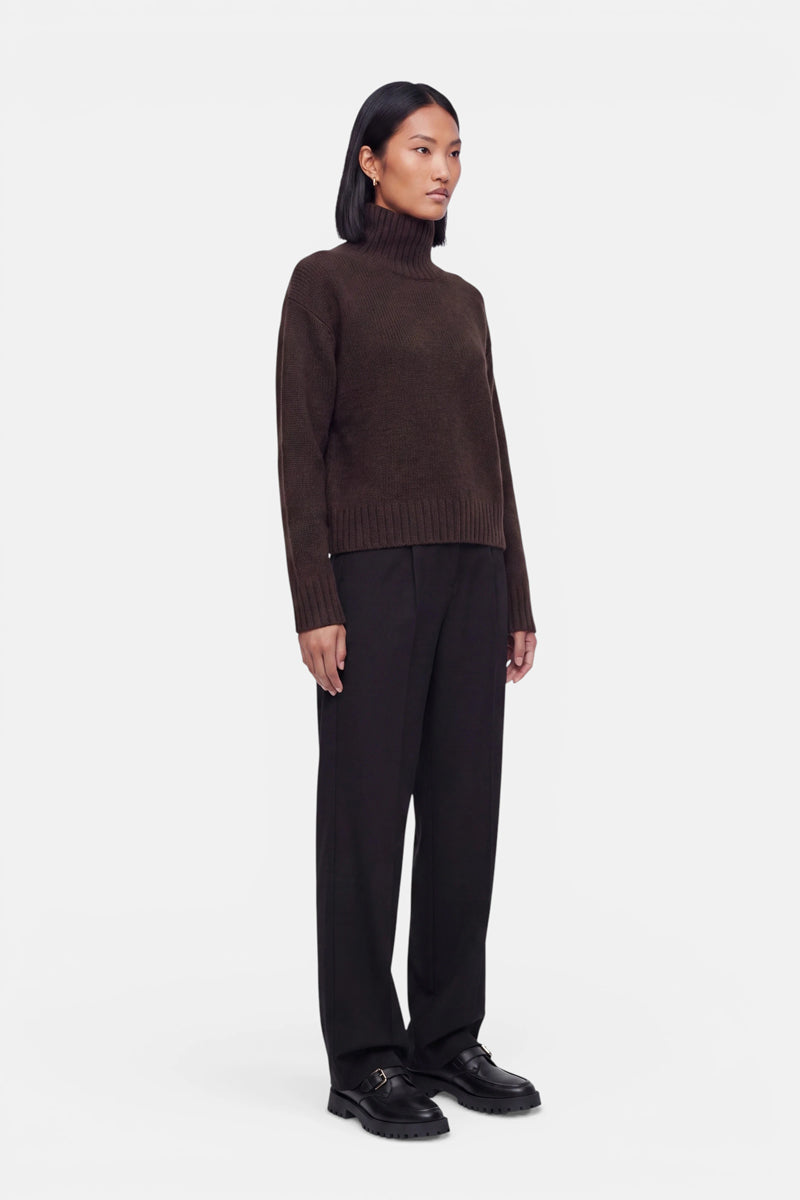 Eine Person steht vor einem schlichten weißen Hintergrund und trägt einen dunkelbraunen Kaschmir-Strickpullover mit Stehkragen, dazu eine weite schwarze Hose und schwarze Schuhe. Die Person hat glattes, schulterlanges schwarzes Haar und einen neutralen Gesichtsausdruck; die Arme hängen entspannt an den Seiten herab.