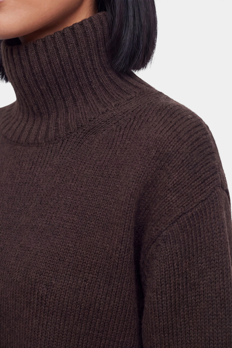 Eine Nahaufnahme einer Person, die einen dicken, gerippten, dunkelbraunen Stehkragen-Kaschmir-Strickpullover trägt. Das Bild hebt die luxuriöse Textur und die detailreiche Strickoptik des Pullovers hervor, wobei das kurze, glatte schwarze Haar der Person über dem hohen Kragen zu sehen ist. Der Hintergrund ist einfarbig und hell.