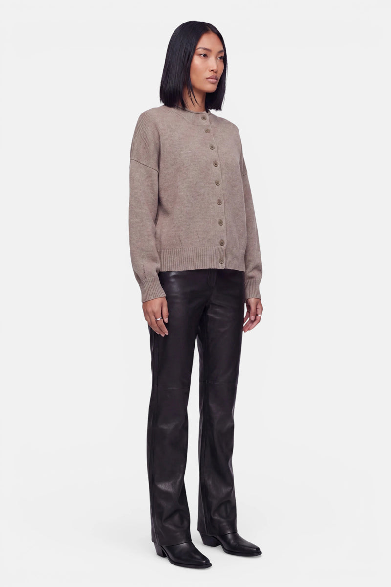 Eine Frau steht leicht nach rechts gewandt und trägt eine taupefarbene Kaschmir-Strickjacke mit langen Ärmeln und Rundhalsausschnitt, dazu eine schwarze Lederhose und schwarze Stiefel. Sie hat glattes, schulterlanges dunkles Haar und einen neutralen Gesichtsausdruck. Der Hintergrund ist schlicht weiß.