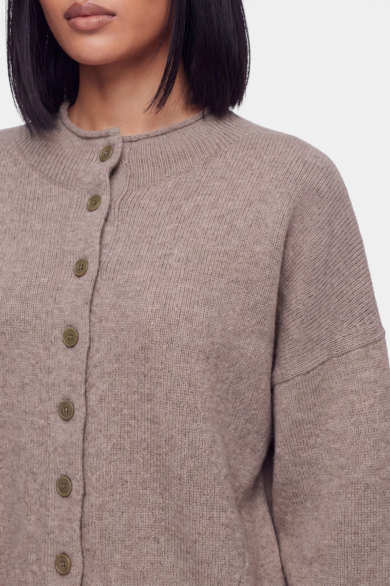 Eine Frau mit glattem, schulterlangem dunklem Haar trägt eine taupefarbene, gerippte Kaschmir-Strickjacke mit Rundhalsausschnitt und großen olivfarbenen Knöpfen. Das Bild ist so beschnitten, dass sie von den Schultern bis zur Taille zu sehen ist, wodurch die Struktur und die Details der Strickjacke – wie beispielsweise die langen Ärmel – vor einem schlichten Hintergrund besonders gut zur Geltung kommen.