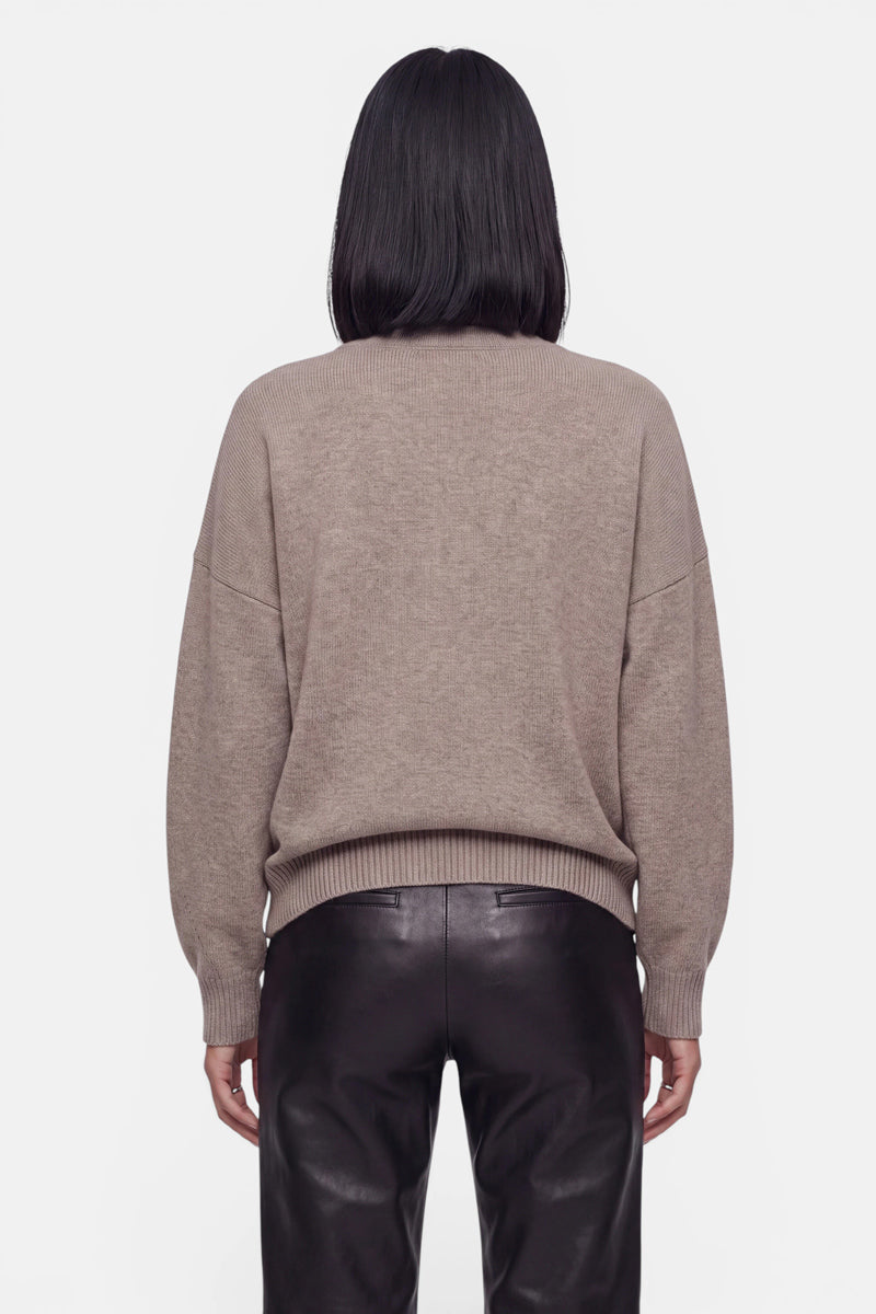 Eine Person steht mit dem Rücken zur Kamera und präsentiert eine Damen-Kaschmir-Strickjacke mit Rundhalsausschnitt, überschnittenen Schultern sowie gerippten Bündchen und Saum. Der taupefarbene Strickpullover wird mit einer schwarzen Lederhose mit hoher Taille kombiniert. Das glatte, schulterlange dunkle Haar hebt sich deutlich vom reinweißen Hintergrund ab.