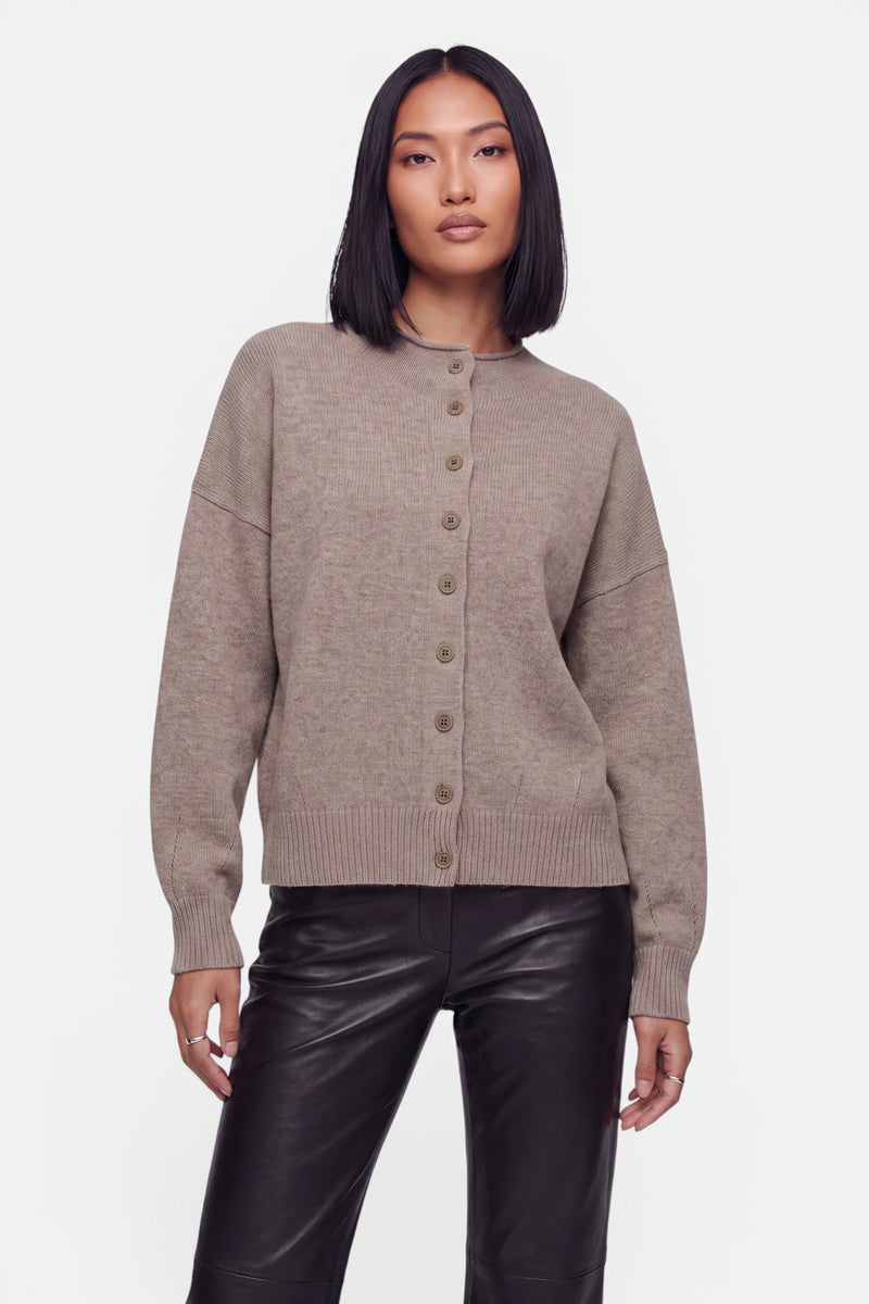 Eine Frau mit glattem, schulterlangem dunklem Haar trägt eine taupefarbene Kaschmir-Strickjacke mit Rundhalsausschnitt, gerippten Bündchen und Saum sowie langen Ärmeln, dazu eine schwarze Lederhose mit hoher Taille; sie steht vor einem schlichten weißen Hintergrund.