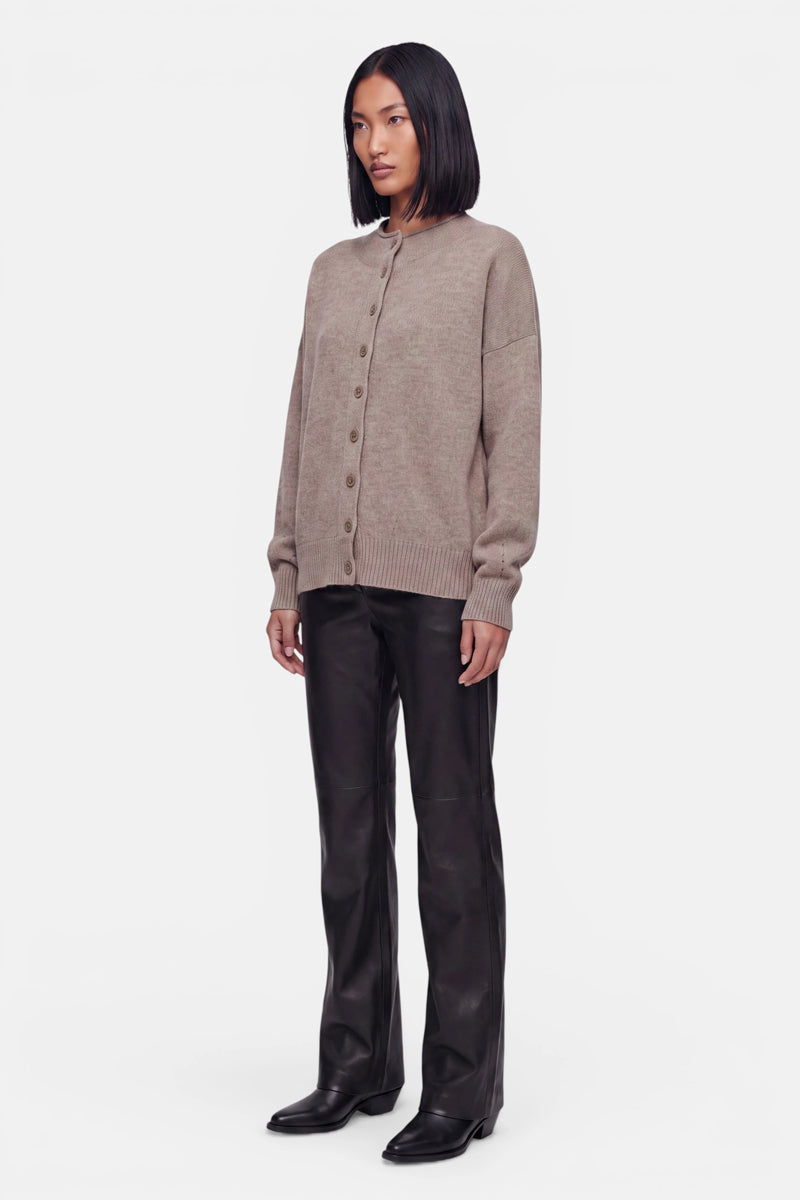 Eine Frau mit glattem, schulterlangem schwarzem Haar trägt eine beige Kaschmir-Strickjacke mit Rundhalsausschnitt und langen Ärmeln, dazu schwarze Lederhosen und schwarze Schuhe. Sie steht vor einem schlichten weißen Hintergrund, blickt leicht nach links und hat einen neutralen Gesichtsausdruck sowie eine entspannte Körperhaltung.