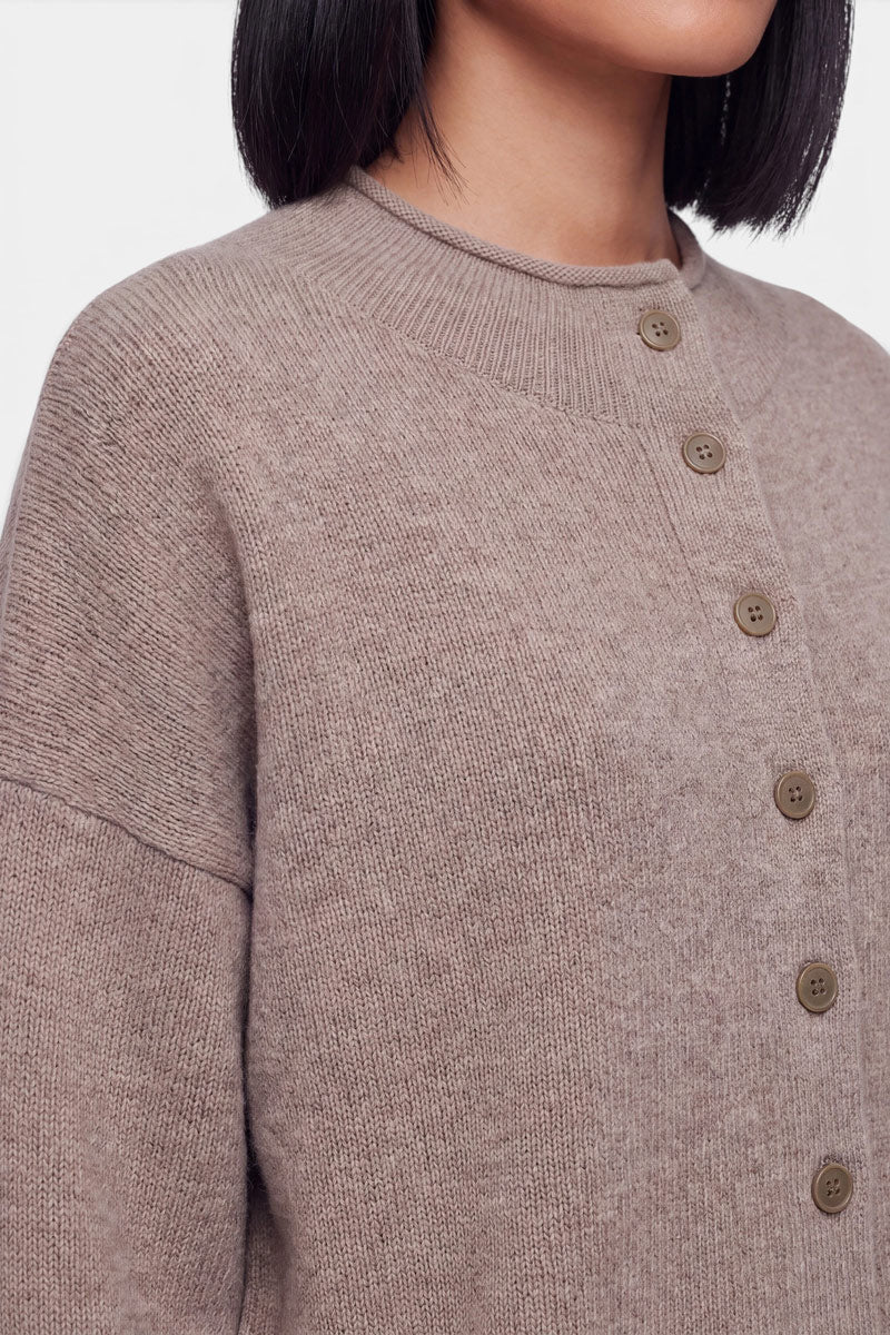 Eine Person mit glattem, dunklem Haar trägt eine hellbraune Kaschmir-Strickjacke mit langen Ärmeln und einem gerippten Kragen. Das Bild ist ab dem Mund abgeschnitten, sodass der Fokus auf der Textur des Kleidungsstücks, dem Rundhalsausschnitt und den Knopfdetails liegt. Der Hintergrund ist einfarbig und hell.
