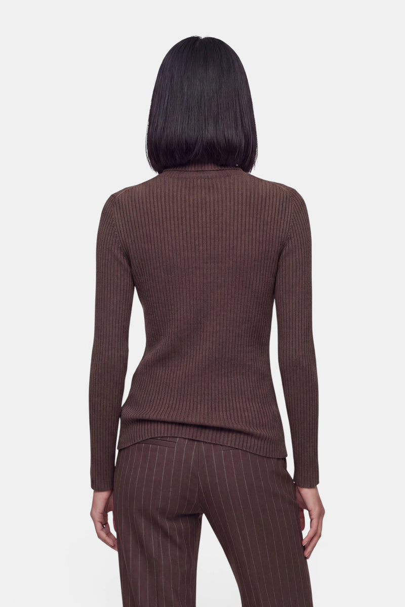 Eine Person mit glattem, schulterlangem, dunklem Haar ist von hinten zu sehen. Sie trägt einen gerippten Pullover in sattem Braun und eine braun gestreifte Hose vor einem schlichten weißen Hintergrund.