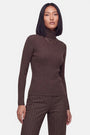 Eine Frau mit glattem, schulterlangem schwarzem Haar trägt einen eng anliegenden, gerippten Strickpullover in Braun und eine dazu passende braune Nadelstreifenhose. Sie steht mit neutralem Gesichtsausdruck nach vorne gerichtet da und trägt eine goldene Halskette und einen goldenen Ring. Schlichter weißer Hintergrund.