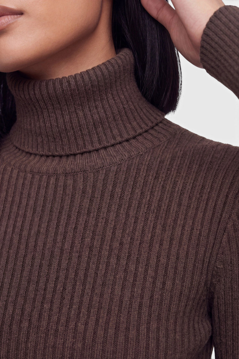Eine Person mit mittlerem Hautton trägt einen gerippten Pullover in einem satten Braunton, der als klassischer Rollkragenpullover gestylt ist. Das Bild hebt die Kinnlinie, den Hals und einen Teil der Schulter hervor, wobei eine Hand zum Kopf erhoben ist. Glattes, dunkles Haar umrahmt die Szene vor einem neutralen Hintergrund und unterstreicht den eleganten Look des Strickpullovers für Damen.