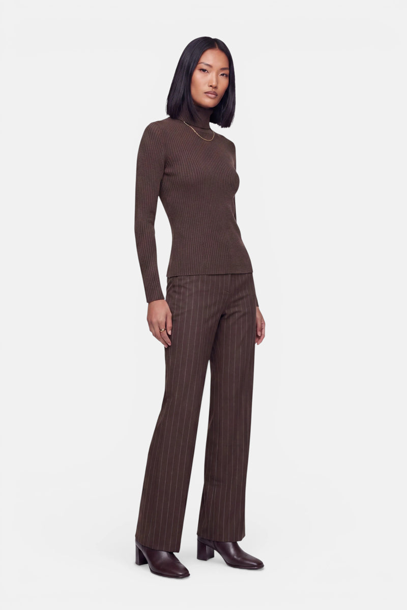 Eine Frau mit glattem schwarzem Haar steht vor einem weißen Hintergrund und trägt einen eng anliegenden braunen Kaschmir-Rollkragenpullover mit gerippter Strickstruktur, eine braune, fein gestreifte Hose mit weitem Bein, Stiefel mit Absatz und eine dünne goldene Halskette. Sie blickt mit ausdruckslosem Blick nach vorne.