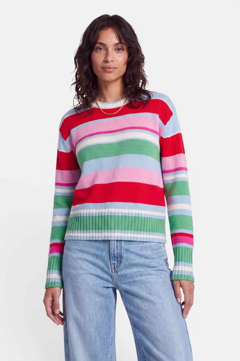 Eine Person, die einen bunten, gestreiften Strickpullover aus einer Woll-Kaschmir-Mischung mit roten, grünen, rosa, blauen und weißen Streifen trägt, steht mit dem Blick nach vorne. Sie hat welliges dunkles Haar, helle Haut und trägt hellblaue Jeans. Der Hintergrund ist einfarbig weiß.