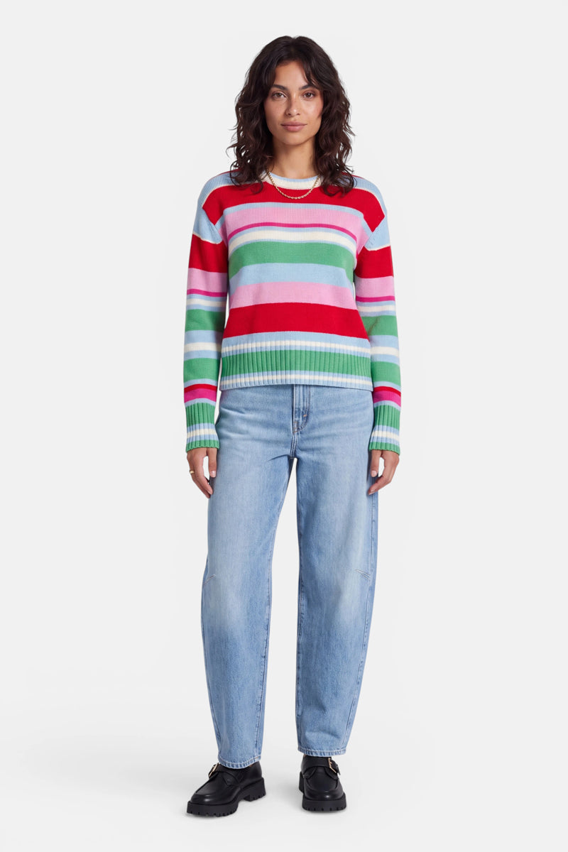 Eine Frau mit welligem braunem Haar trägt einen gestreiften Strickpullover in Rot-, Rosa-, Weiß- und Grüntönen, dazu hellblaue Jeans und schwarze Schuhe. Der farbenfrohe Pullover aus einer weichen Wolle-Kaschmir-Mischung fällt besonders ins Auge, während sie mit vor dem schlichten weißen Hintergrund stehend die Hände entspannt an den Seiten hält.