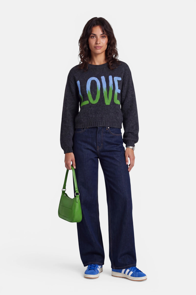 Eine Frau steht vor weißem Hintergrund und trägt einen dunklen Strickpullover aus Merino-Alpaka mit einer „LOVE“-Intarsie in Grün und Blau, eine weite dunkle Jeans, blaue Adidas-Sneaker und hält eine kleine grüne Handtasche in der Hand. Sie hat mittellanges, welliges braunes Haar und einen neutralen Gesichtsausdruck.