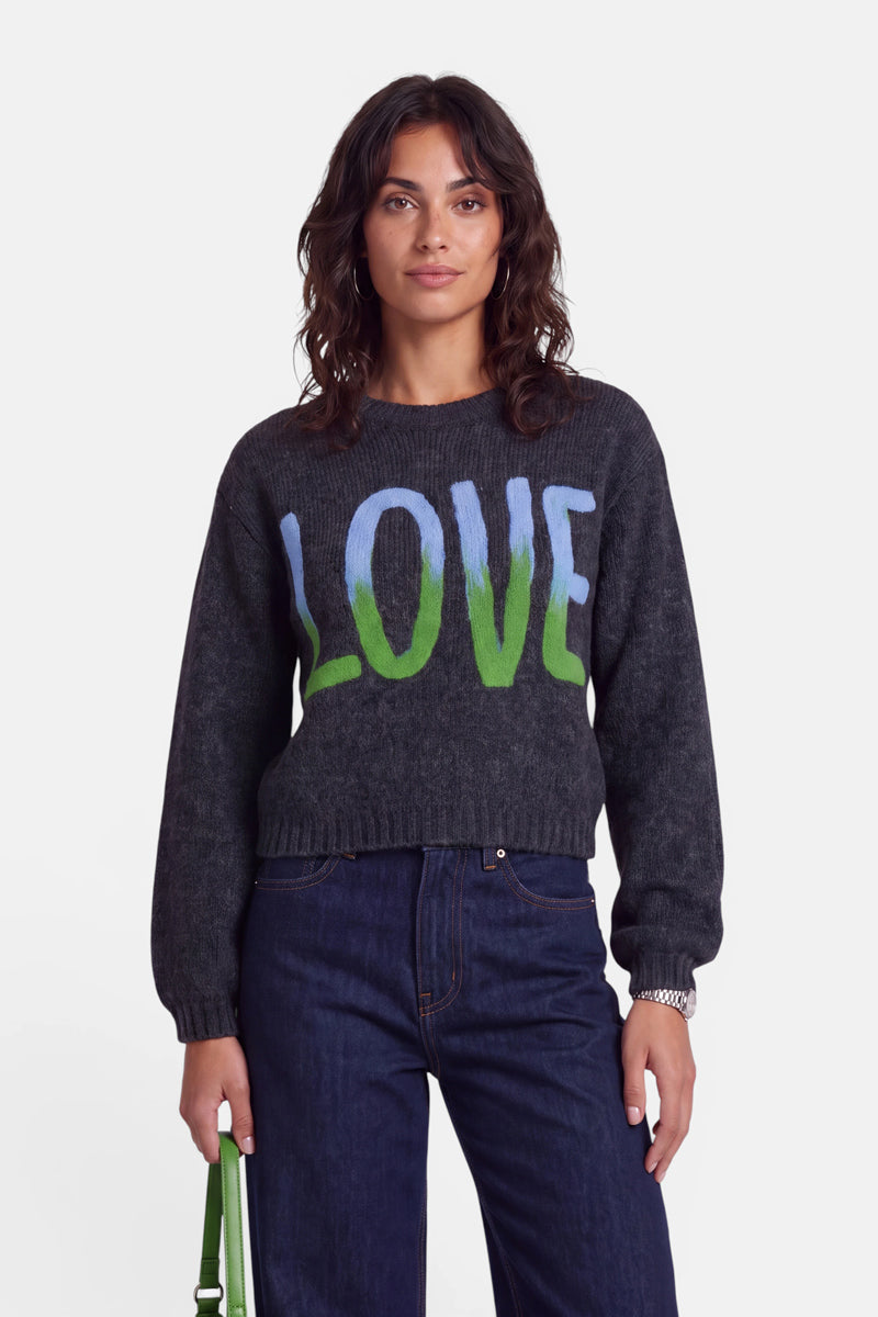 Eine Frau mit welligem braunem Haar trägt einen dunkelgrauen Strickpullover aus Merino-Alpaka mit der Aufschrift „LOVE“ in großen blauen und grünen Buchstaben auf der Vorderseite. Der Pullover hat einen klassischen Rundhalsausschnitt. Sie trägt dunkelblaue Jeans, eine silberne Armbanduhr und hält eine grüne Handtasche in der Hand. Sie steht vor einem schlichten weißen Hintergrund, blickt nach vorne und lächelt leicht.