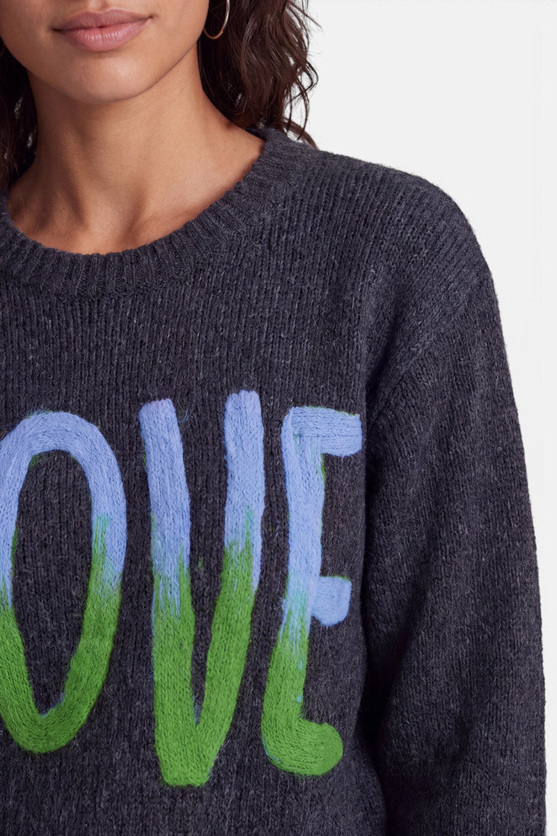 Eine Frau in einem dunklen Strickpullover aus Merino-Alpaka mit Rundhalsausschnitt, verziert mit großen, im Malstil gestalteten Buchstaben O, V und E in Blau und Grün als „Love“-Intarsie. Nur ihre Lippen, ihr Kinn und ein Teil ihres lockigen Haares sind zu sehen. Sie trägt Creolen und steht vor einem schlichten, hellen Hintergrund.