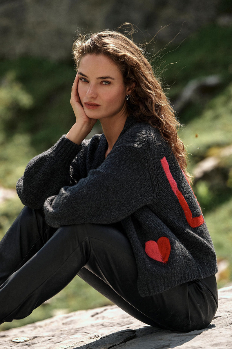 Eine Frau mit welligem braunem Haar sitzt draußen auf einem Felsen und stützt ihr Gesicht auf die Hand. Sie trägt eine dunkle, handgestrickte Strickjacke aus einer Merino-Alpaka-Mischung mit roten Herz-Intarsien und rechteckigen Mustern, dazu eine schwarze Hose. Der Hintergrund ist grasbewachsen und sonnenbeschienen, was auf eine natürliche Umgebung hindeutet.