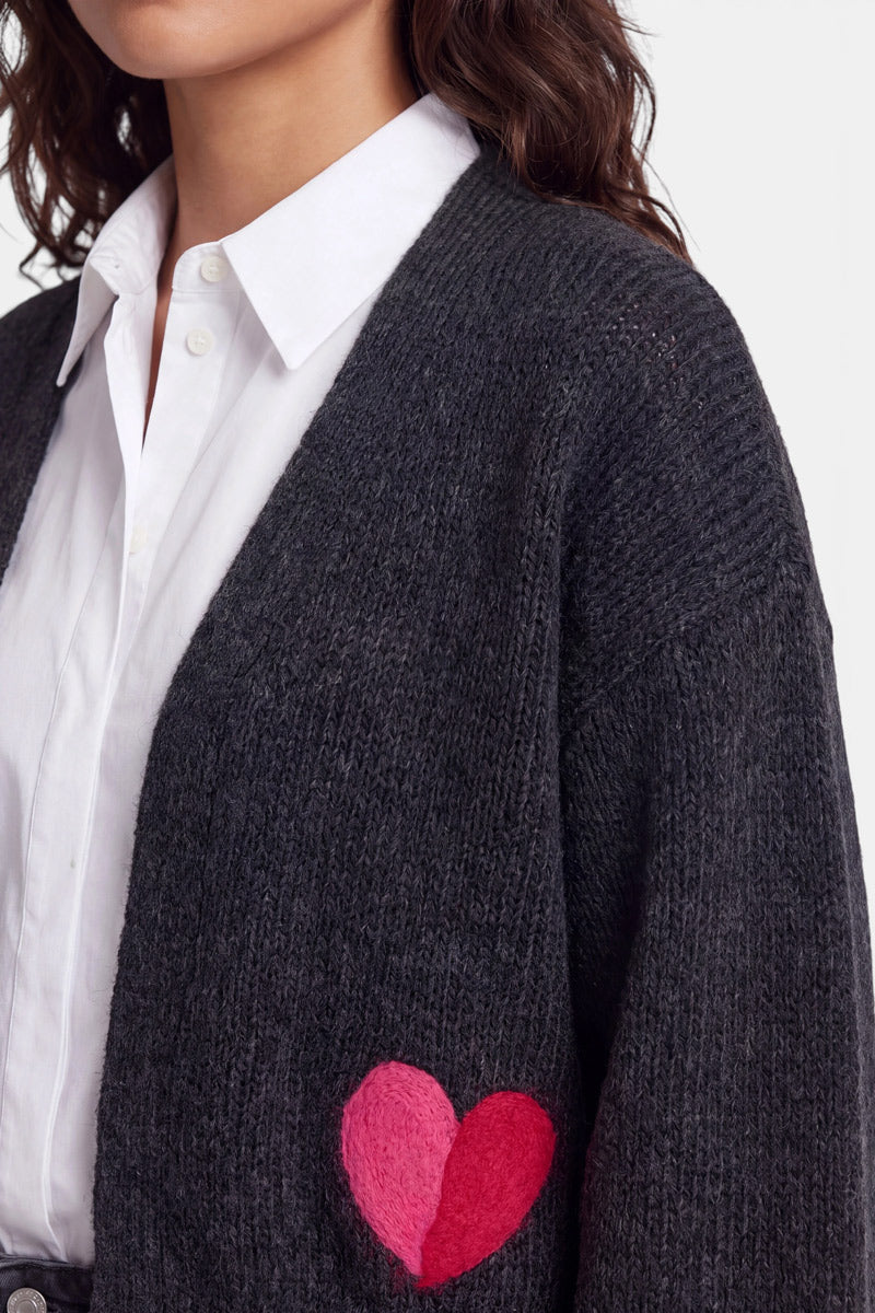 Eine Frau mit welligem braunem Haar trägt ein weißes Hemd unter einer dunkelgrauen Strickjacke aus Merino- und Alpaka-Wolle, die am Ärmel mit einem rosa-roten Herz-Intarsien-Aufnäher verziert ist. Nur ihr Oberkörper ist zu sehen, der Hintergrund ist einfarbig hellgrau.